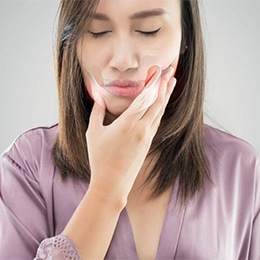 Woman in purple shirt touching her cheek, appearing to experience tooth or jaw pain, gray background.