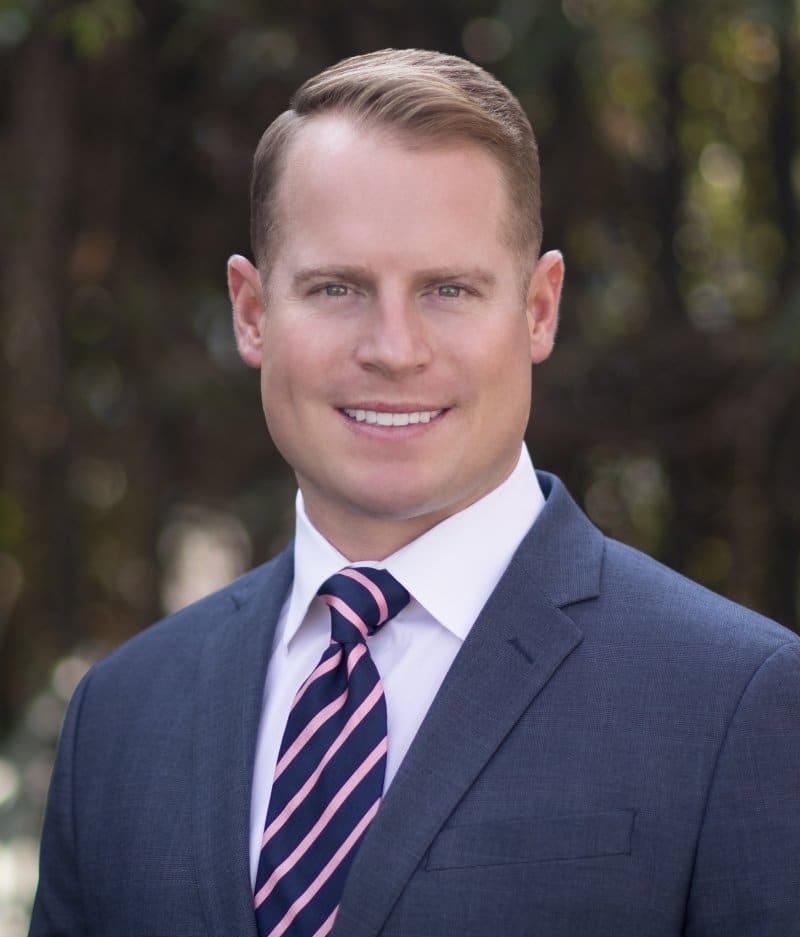 Professional headshot of male dentist smiling outdoors, wearing blue suit and striped tie.