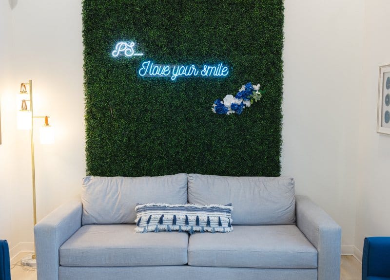 Modern dental office waiting area with gray couch beneath neon "Prosper Smile" sign and green plant wall.