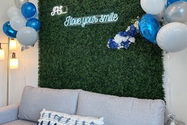 Dental office photo backdrop with artificial grass wall, blue balloons, and "Blue you smile" text decoration.