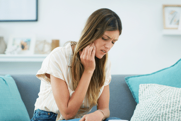 Woman sitting on turquoise couch holding cheek, appearing to experience tooth or jaw pain indoors.
