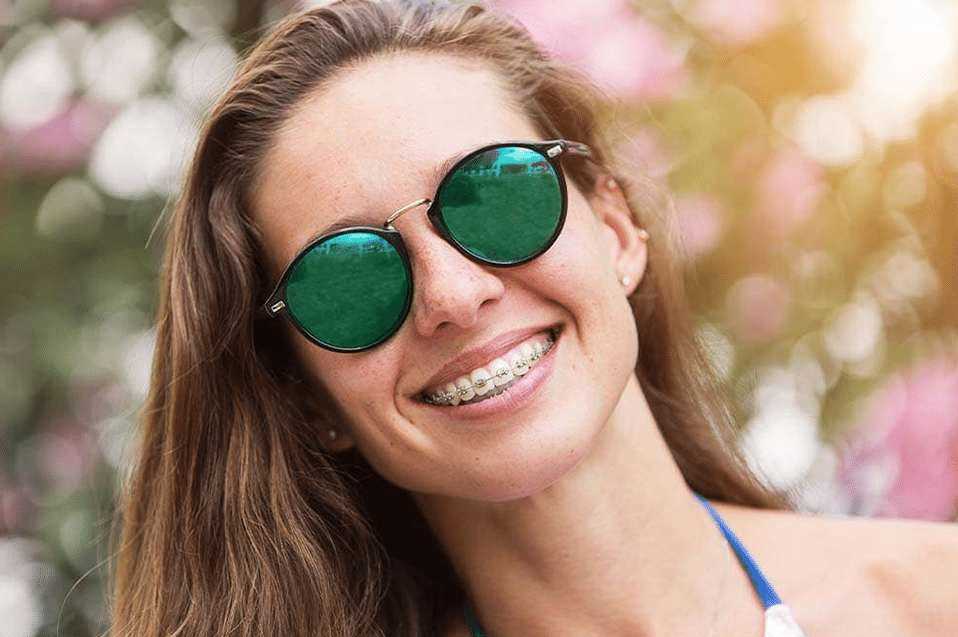 Woman wearing green-tinted sunglasses smiling outdoors with blurred floral background.
