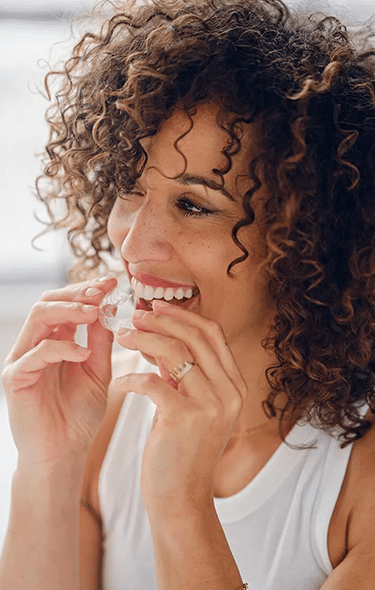 Woman with curly brown hair smiling while flossing teeth in bright bathroom setting.