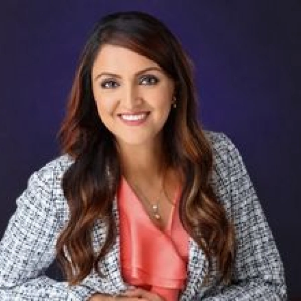 Professional studio headshot of Dr. Jina wearing a tweed blazer and coral top against a dark blue background.
